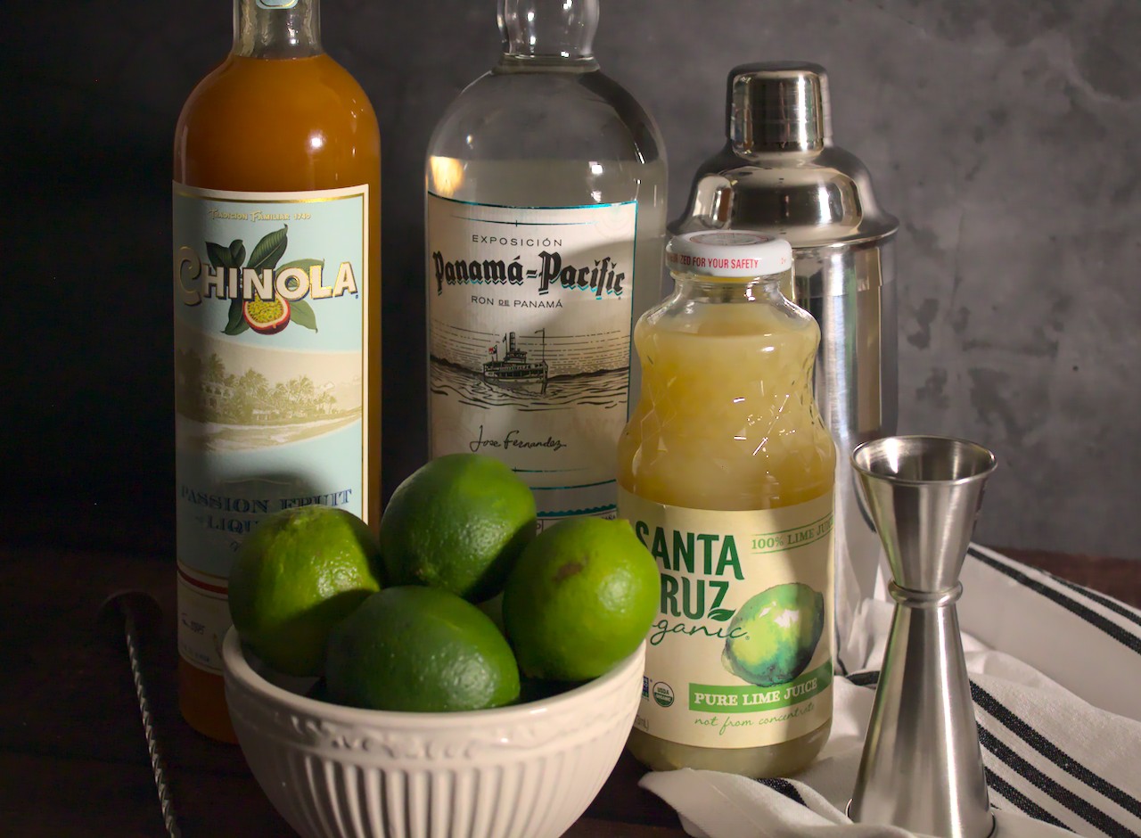 Photo of limes, lime juice, passion fruit liqueur, and rum next to a cocktail shaker