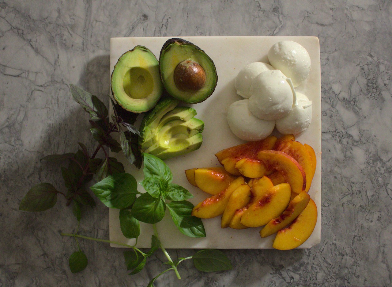 Overhead shot of sliced nectarines, avocado, buffalo mozzarella, and basil on a cutting board